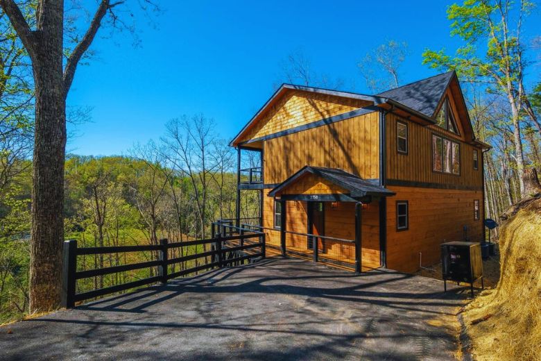 Modern wood cabin with black trim and a pitched roof, set on a paved driveway with a wooden fence, surrounded by trees and overlooking a forested valley under a clear blue sky.